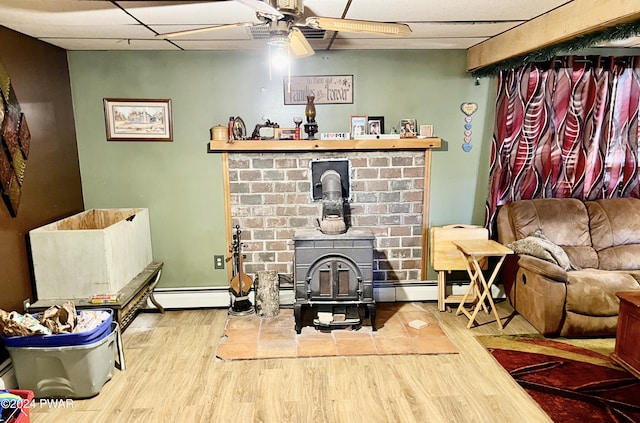 living room with a drop ceiling, a wood stove, hardwood / wood-style flooring, ceiling fan, and baseboard heating