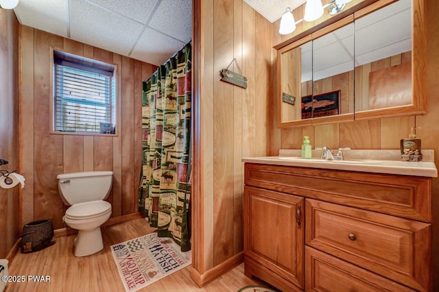 bathroom featuring toilet, wood walls, vanity, wood finished floors, and a drop ceiling