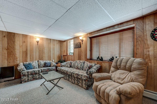 carpeted living area with wooden walls, baseboard heating, and a drop ceiling