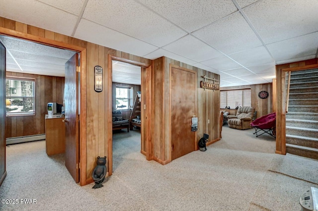 hallway featuring a paneled ceiling, wooden walls, carpet flooring, stairs, and baseboard heating