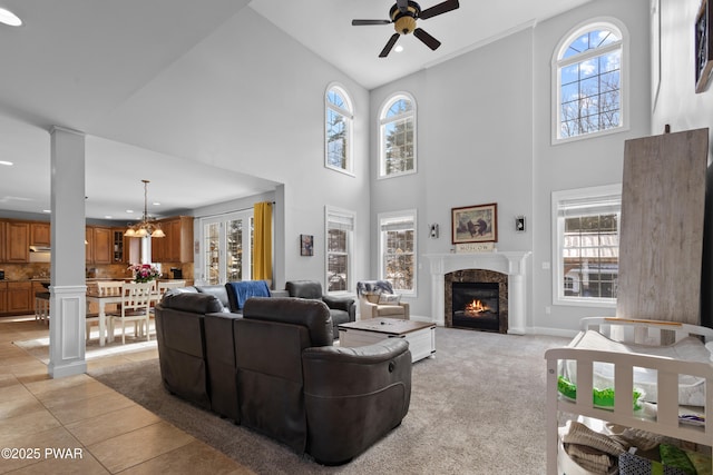 living area with light tile patterned floors, a lit fireplace, ceiling fan with notable chandelier, and decorative columns