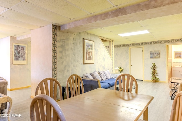 dining space featuring light wood finished floors, wallpapered walls, baseboards, and a drop ceiling