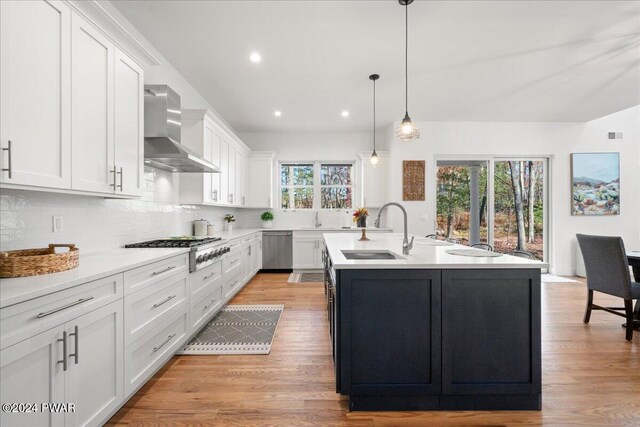 kitchen featuring appliances with stainless steel finishes, wall chimney exhaust hood, sink, white cabinets, and an island with sink