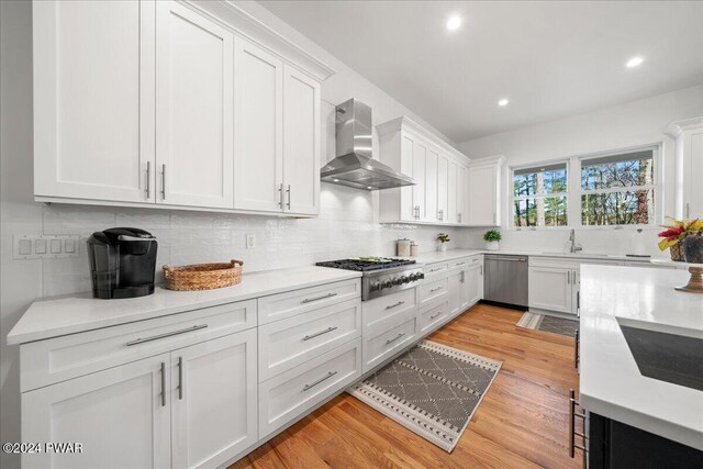 kitchen with white cabinets, wall chimney exhaust hood, appliances with stainless steel finishes, and light hardwood / wood-style flooring