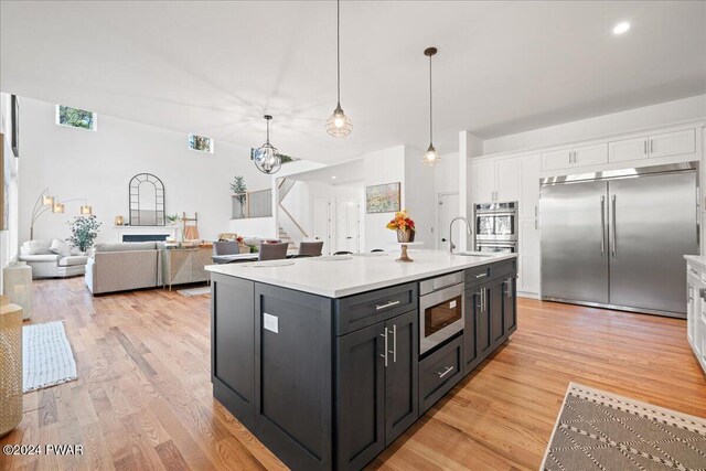 kitchen featuring built in appliances, decorative light fixtures, light hardwood / wood-style floors, white cabinetry, and an island with sink