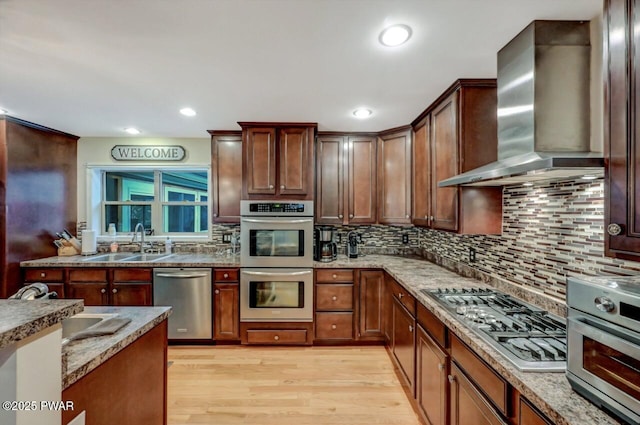 kitchen with light wood-style flooring, a sink, appliances with stainless steel finishes, wall chimney exhaust hood, and tasteful backsplash