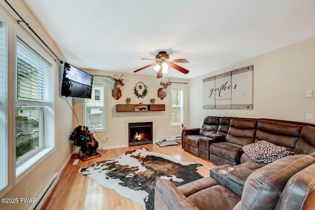 living area featuring a baseboard radiator, a warm lit fireplace, a healthy amount of sunlight, and wood finished floors