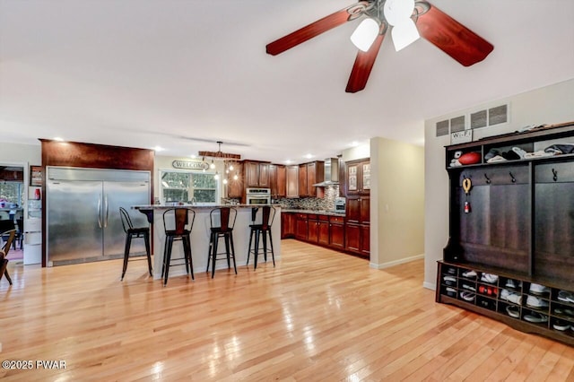 kitchen featuring a breakfast bar, appliances with stainless steel finishes, wall chimney range hood, backsplash, and light wood finished floors