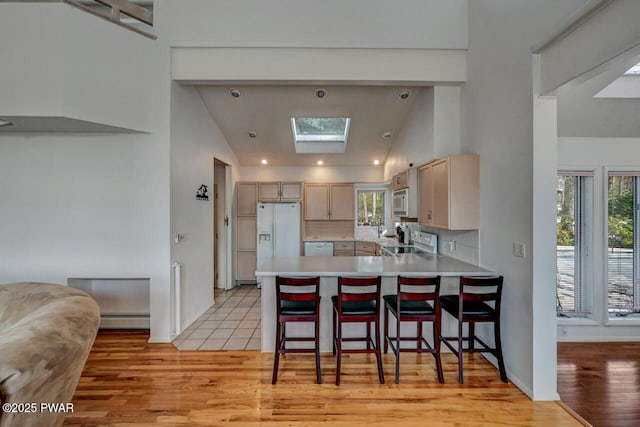 kitchen featuring a breakfast bar, a baseboard radiator, kitchen peninsula, light brown cabinets, and white appliances