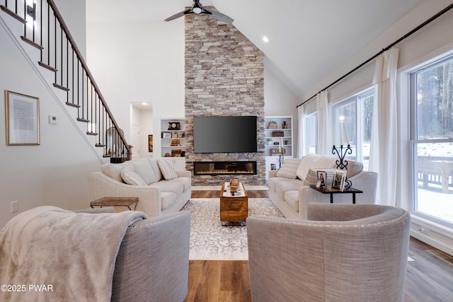 living room featuring a ceiling fan, wood finished floors, stairs, a fireplace, and high vaulted ceiling