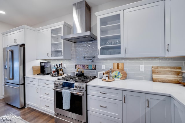 kitchen featuring stainless steel appliances, decorative backsplash, dark wood-type flooring, glass insert cabinets, and wall chimney range hood