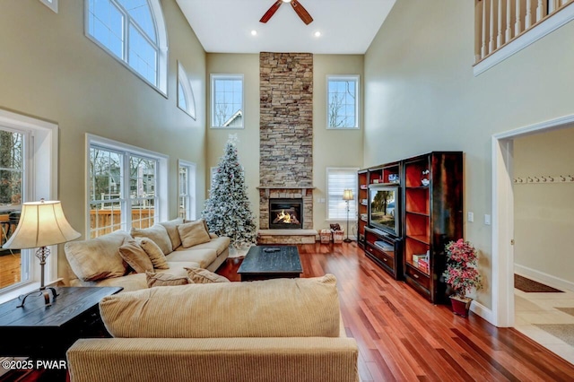 living area with ceiling fan, a stone fireplace, wood finished floors, a towering ceiling, and baseboards