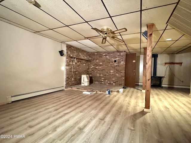 basement with ceiling fan, a drop ceiling, brick wall, a baseboard heating unit, and light wood-type flooring