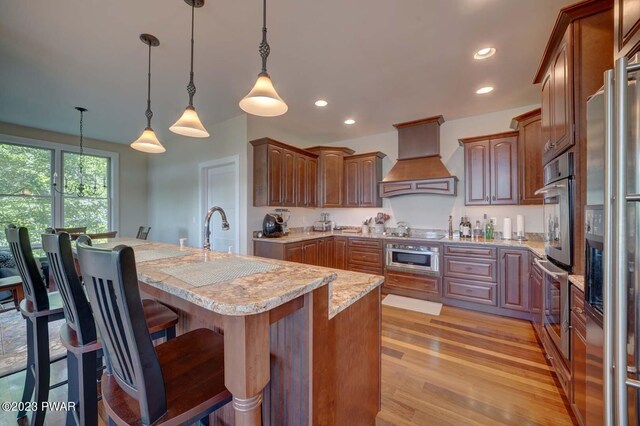 kitchen featuring pendant lighting, a breakfast bar, a kitchen island with sink, light hardwood / wood-style flooring, and custom range hood