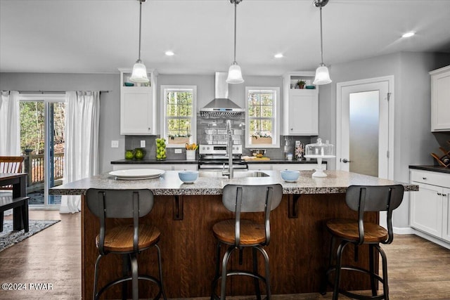 kitchen with a kitchen bar, white cabinetry, an island with sink, pendant lighting, and wall chimney range hood