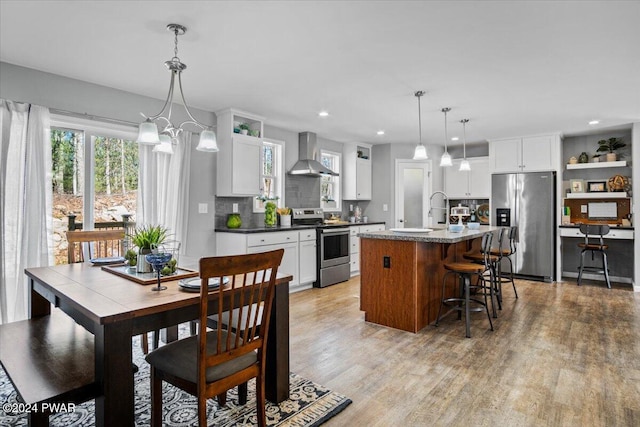 kitchen featuring appliances with stainless steel finishes, white cabinetry, hanging light fixtures, a kitchen island with sink, and wall chimney exhaust hood