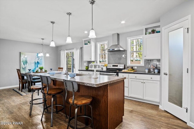 kitchen featuring pendant lighting, white cabinetry, wall chimney range hood, a center island with sink, and stainless steel electric range