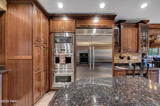 kitchen with brown cabinetry, dark stone countertops, built in appliances, and tasteful backsplash