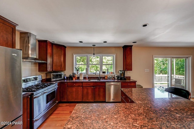 kitchen featuring dark stone counters, wall chimney range hood, sink, light wood-type flooring, and appliances with stainless steel finishes