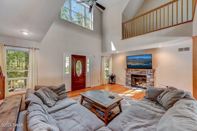 living room featuring ceiling fan, a stone fireplace, a towering ceiling, and light wood-type flooring