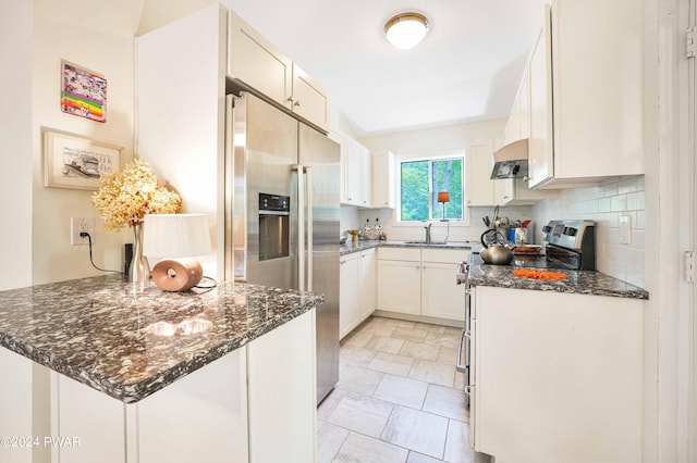 kitchen featuring appliances with stainless steel finishes, tasteful backsplash, dark stone counters, white cabinetry, and range hood