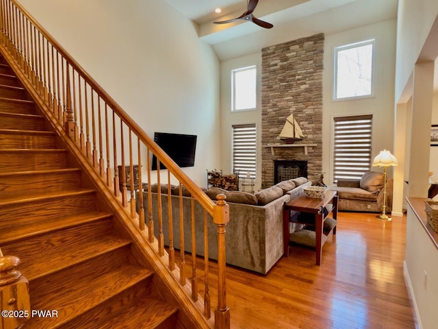 living area with a stone fireplace, wood finished floors, a towering ceiling, a ceiling fan, and stairs