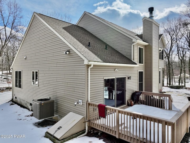 snow covered property featuring central air condition unit, a wooden deck, a chimney, and roof with shingles