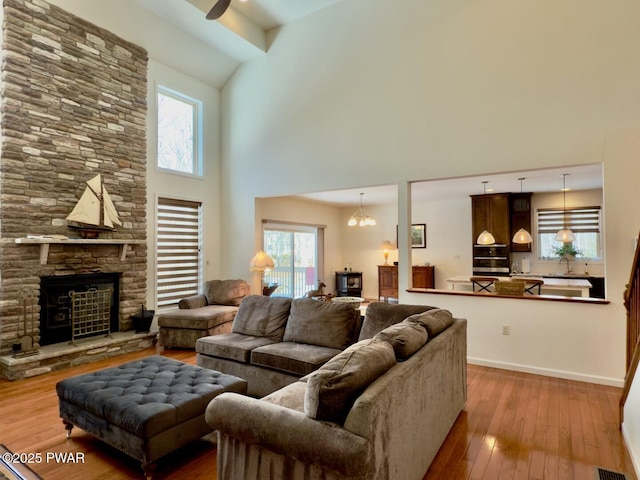 living area with a towering ceiling, hardwood / wood-style floors, a stone fireplace, baseboards, and ceiling fan with notable chandelier