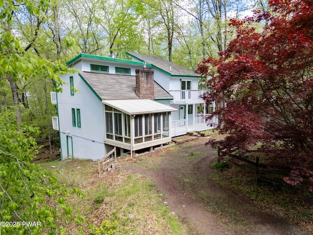 back of property with a balcony, roof with shingles, a sunroom, dirt driveway, and a chimney