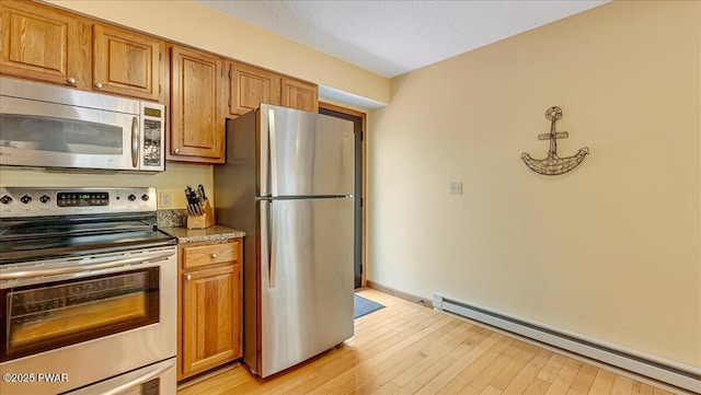 kitchen with appliances with stainless steel finishes, baseboard heating, light wood finished floors, brown cabinetry, and dark stone countertops