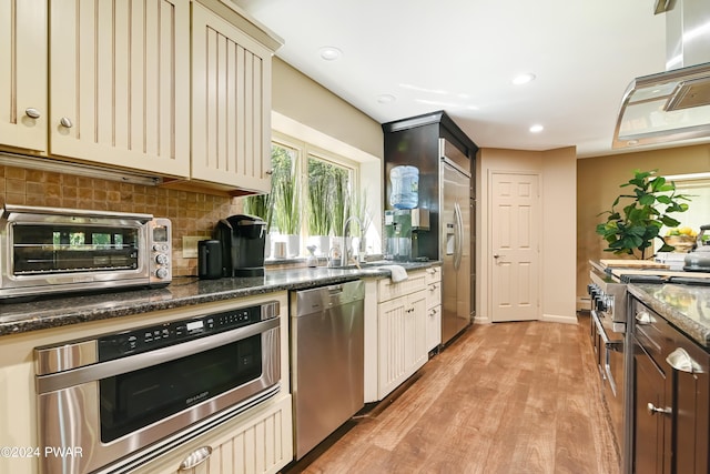 kitchen with appliances with stainless steel finishes, backsplash, dark stone countertops, cream cabinetry, and light hardwood / wood-style floors