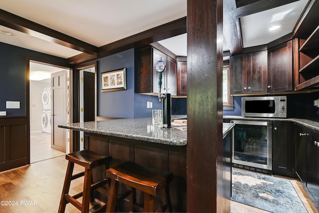 kitchen featuring beverage cooler, stacked washer / dryer, dark stone counters, light hardwood / wood-style floors, and a breakfast bar area