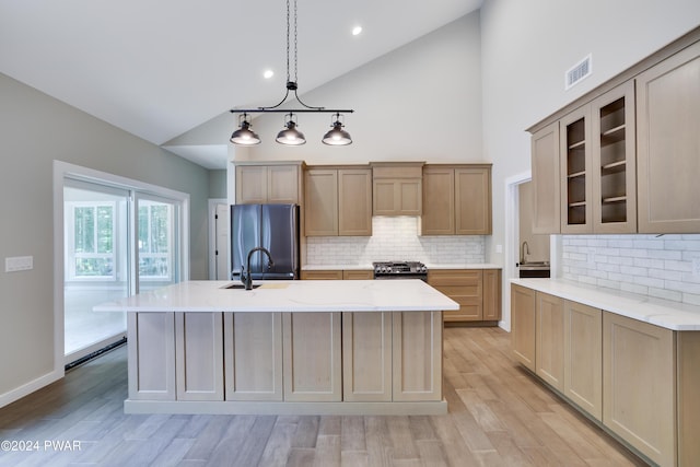 kitchen with backsplash, light stone countertops, an island with sink, light hardwood / wood-style floors, and stainless steel refrigerator