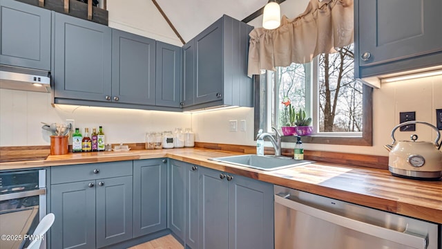 kitchen with wooden counters, blue cabinets, under cabinet range hood, a sink, and stainless steel appliances