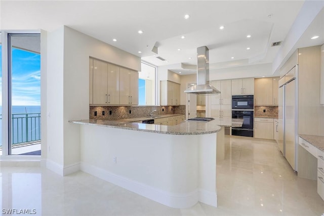 kitchen with light stone counters, dobule oven black, island range hood, a peninsula, and a water view