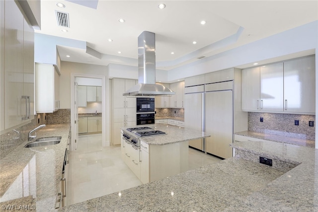 kitchen with tasteful backsplash, light stone counters, paneled built in fridge, a tray ceiling, and island range hood