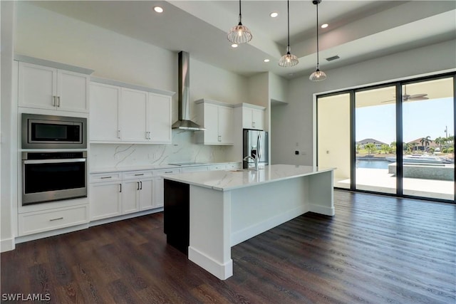 kitchen featuring white cabinets, a center island with sink, hanging light fixtures, wall chimney exhaust hood, and stainless steel appliances