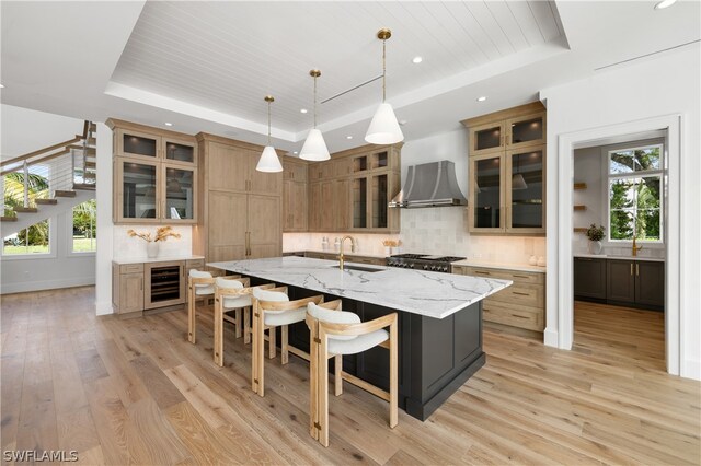 kitchen with a raised ceiling, wall chimney range hood, sink, and a large island with sink