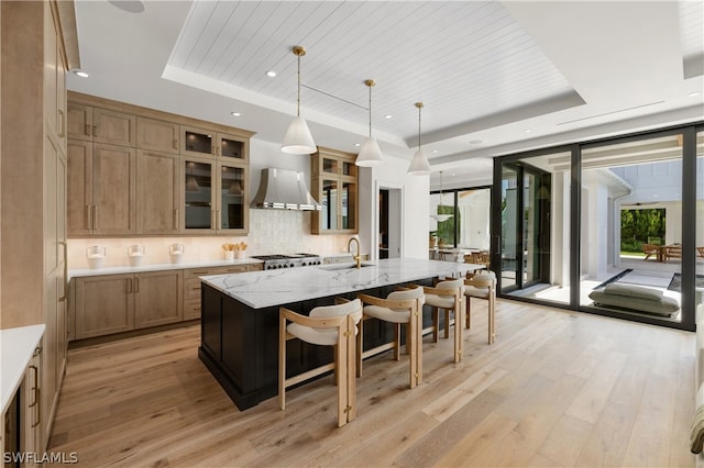 kitchen featuring light hardwood / wood-style flooring, wall chimney range hood, an island with sink, and a tray ceiling