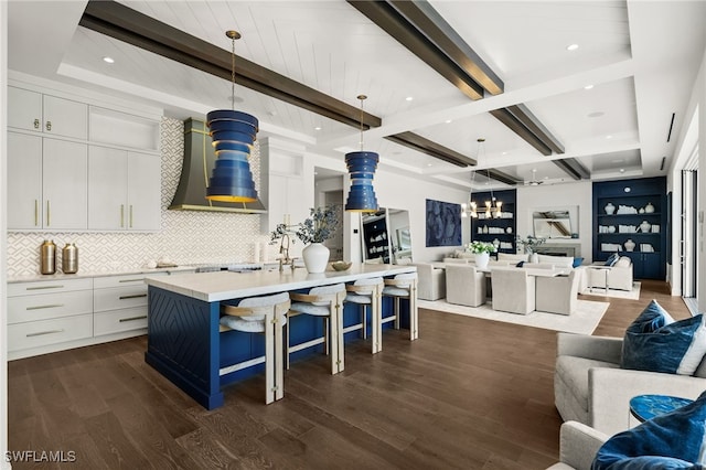 kitchen featuring a kitchen island with sink, wall chimney range hood, hanging light fixtures, dark hardwood / wood-style floors, and beam ceiling