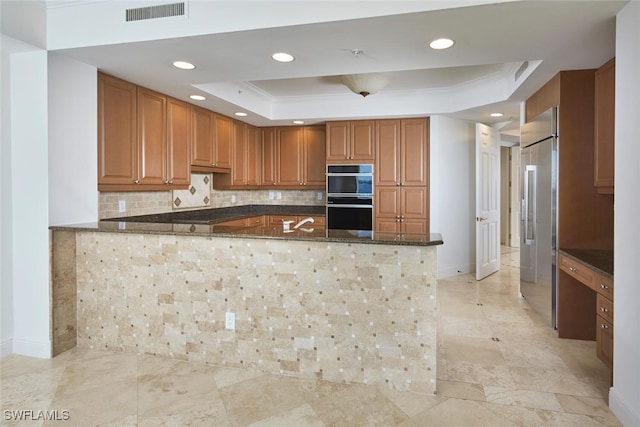 kitchen featuring dark stone counters, a tray ceiling, kitchen peninsula, black double oven, and ornamental molding