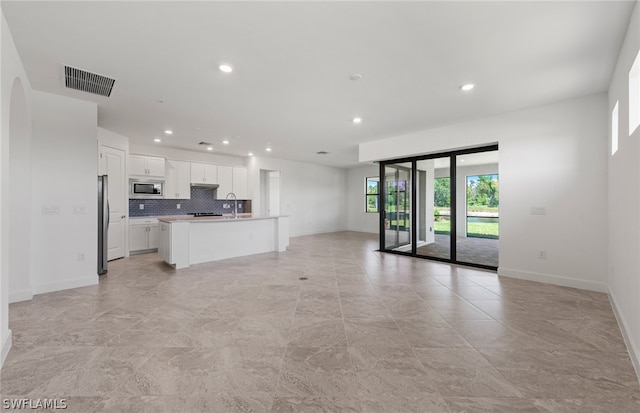 kitchen with stainless steel appliances, an island with sink, white cabinets, backsplash, and light tile flooring