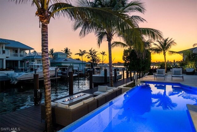 pool at dusk with a boat dock, a water view, and an outdoor fire pit