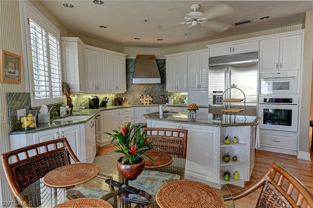 kitchen with ceiling fan, stainless steel built in fridge, wall chimney exhaust hood, dark stone countertops, and a center island