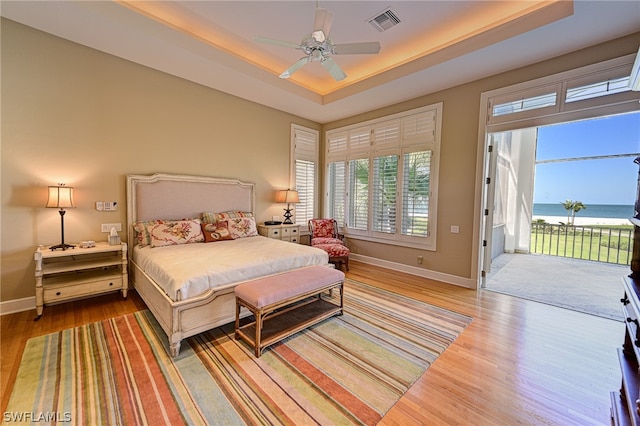 bedroom featuring a water view, light hardwood / wood-style flooring, a tray ceiling, and access to exterior