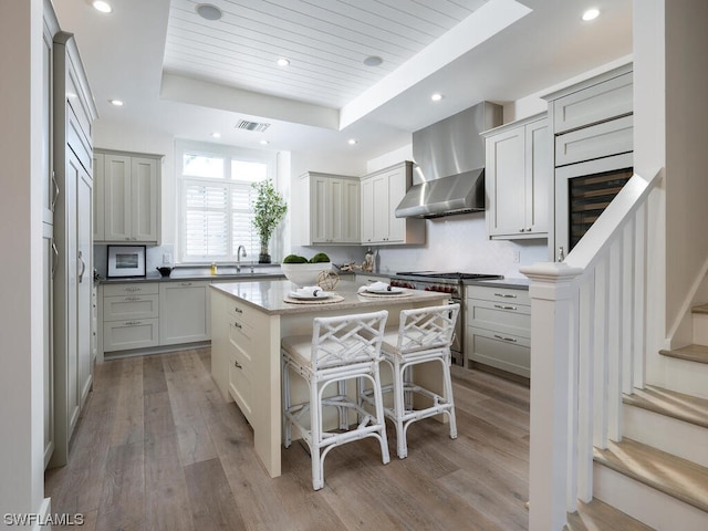 kitchen with a raised ceiling, wall chimney exhaust hood, a kitchen island, and light hardwood / wood-style floors