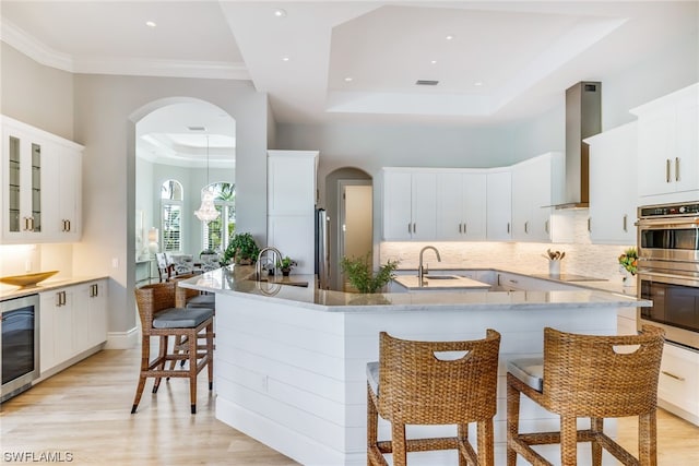 kitchen with a breakfast bar, a raised ceiling, wall chimney exhaust hood, light wood-type flooring, and beverage cooler