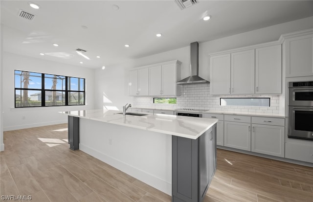 kitchen with wall chimney exhaust hood, a kitchen island with sink, double oven, backsplash, and sink