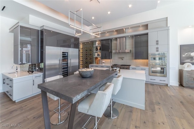 kitchen with sink, a kitchen island with sink, stainless steel appliances, light hardwood / wood-style floors, and wall chimney range hood