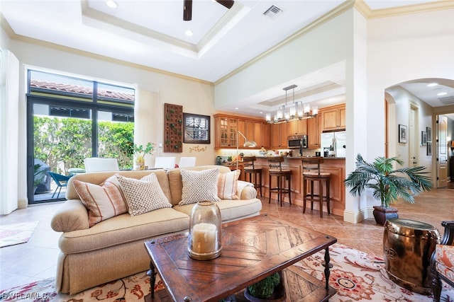 tiled living room with crown molding, a towering ceiling, a raised ceiling, and ceiling fan with notable chandelier